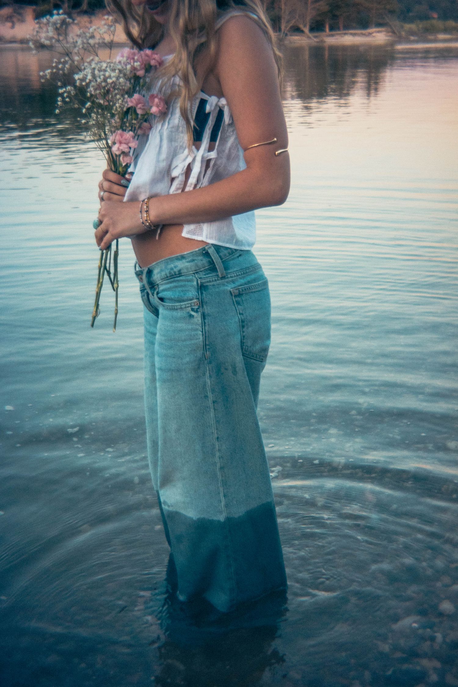Woman in white crop top and wet denim maxi skirt standing in water holding wildflowers.