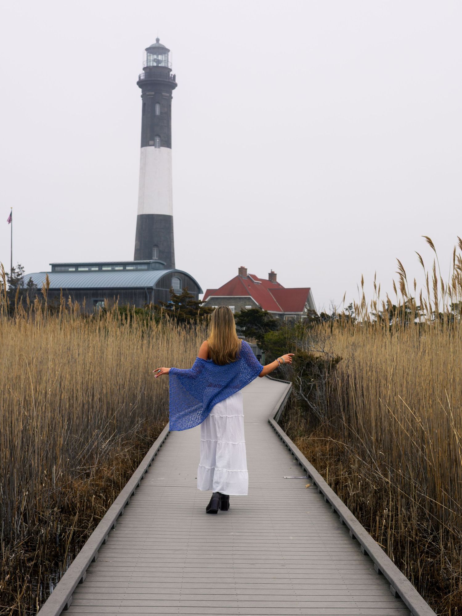 Woman in a white skirt and blue shawl walks a wooden boardwalk through tall marsh grass toward a black and white striped lighthouse.