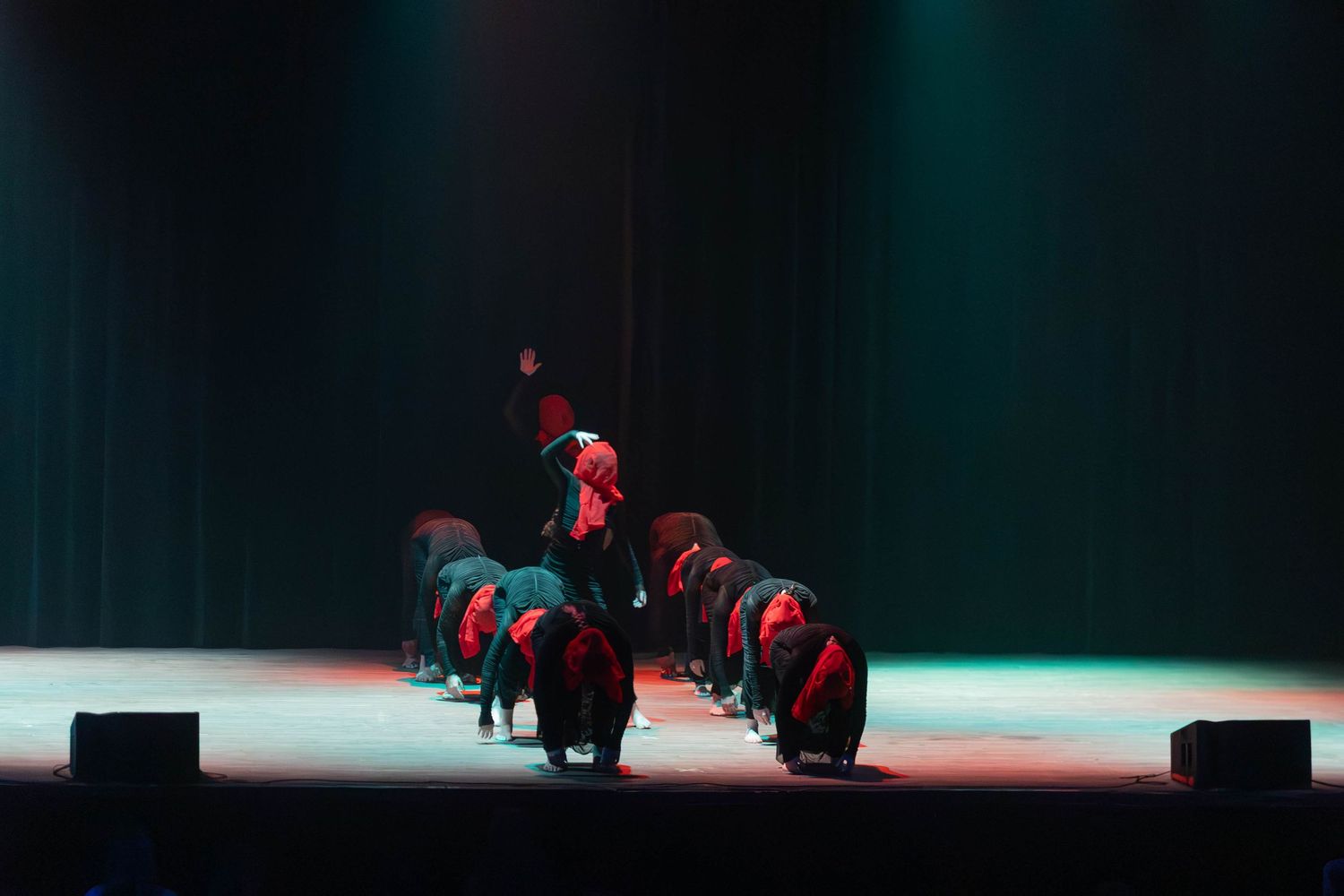 Performers in red and black costumes bow on a dramatic stage with colorful spotlights during a theatrical dance performance.