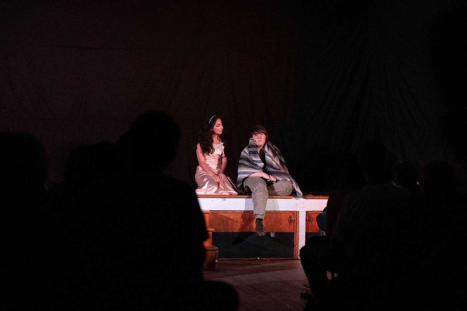 Two actors perform an intimate scene on a white table on a darkened theater stage.