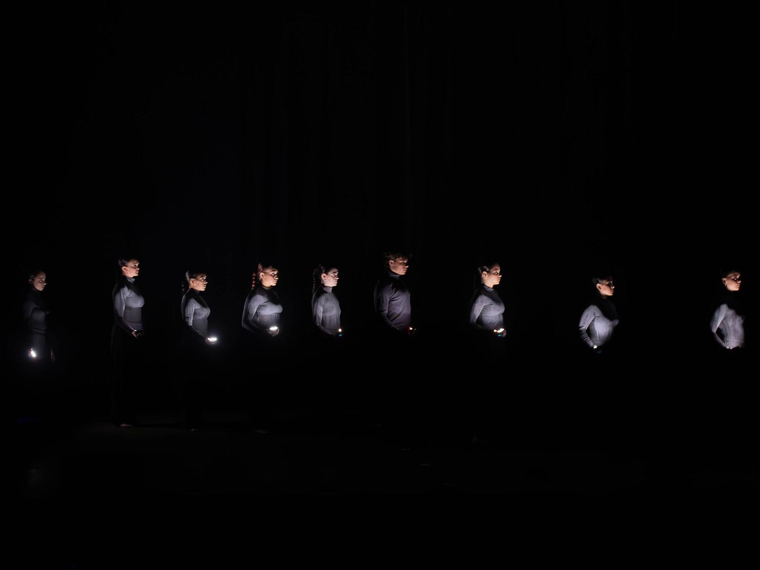 A line of performers in gray uniforms stand illuminated on a dark stage during a dramatic dance performance.