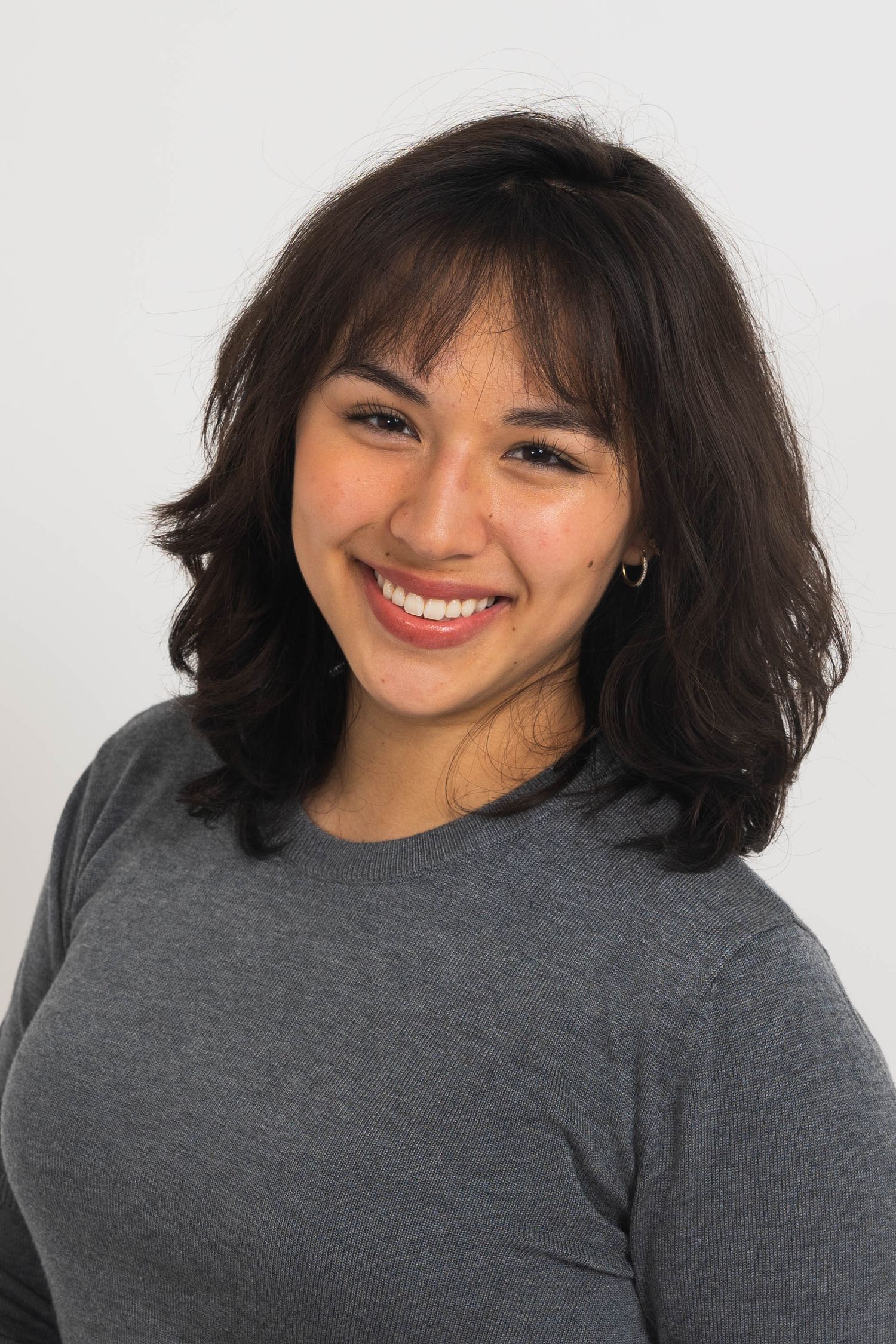 Smiling young woman with shoulder-length wavy dark hair and bangs wearing a gray sweater in front of a plain light studio background.