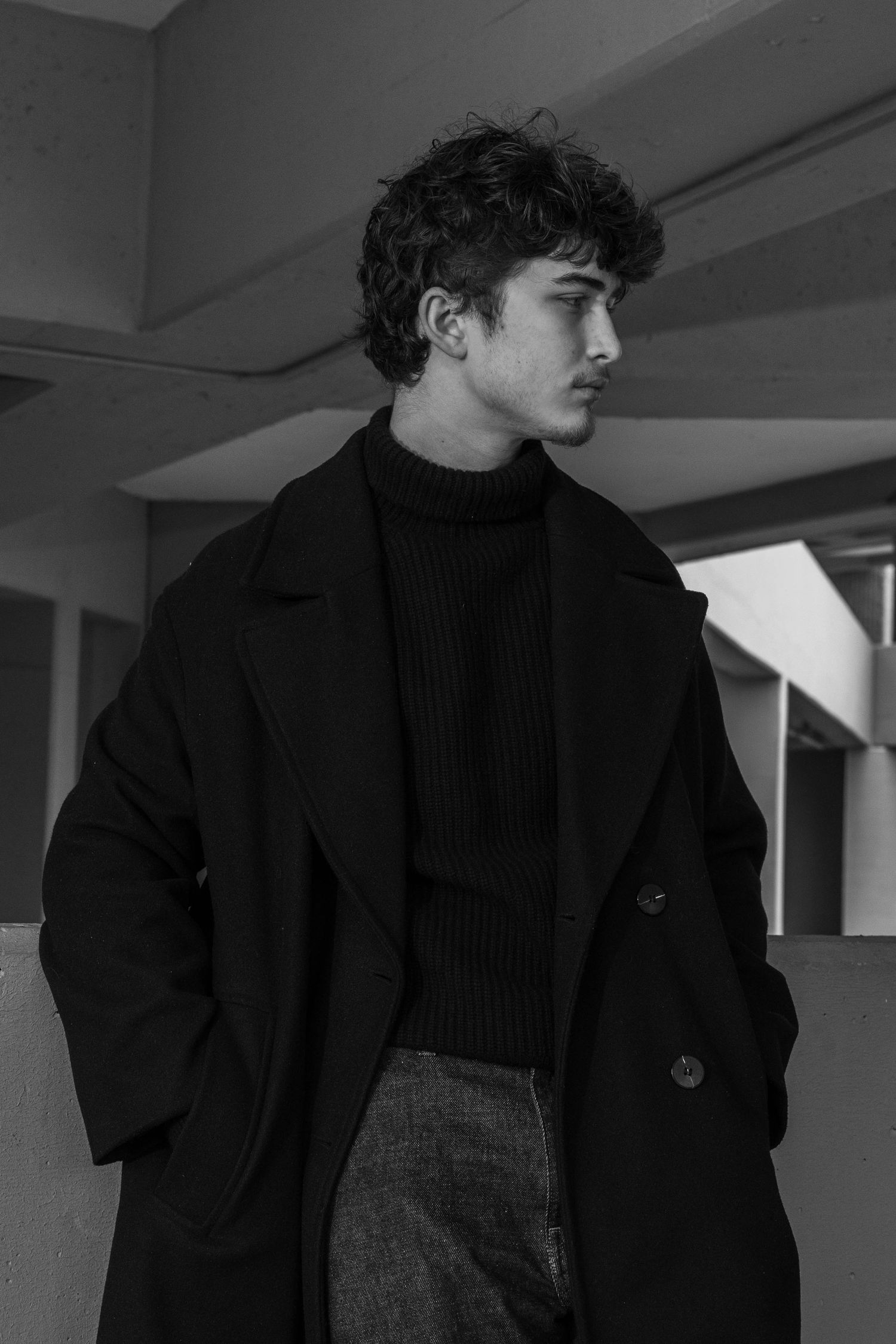 Black and white portrait of a young man with curly hair in a dark overcoat and ribbed turtleneck, standing in profile in a concrete space.