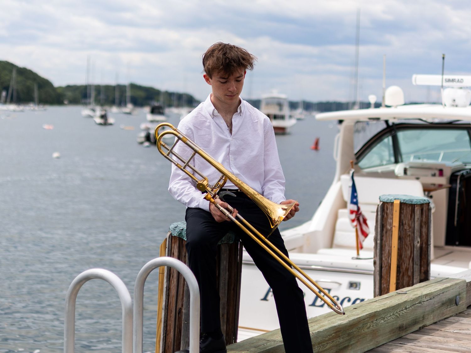 Young musician playing trombone on a marina dock with boats and calm water in the background.