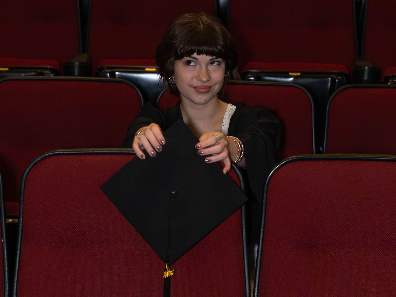 Young woman in auditorium holding graduation cap, celebrating academic achievement at commencement ceremony.