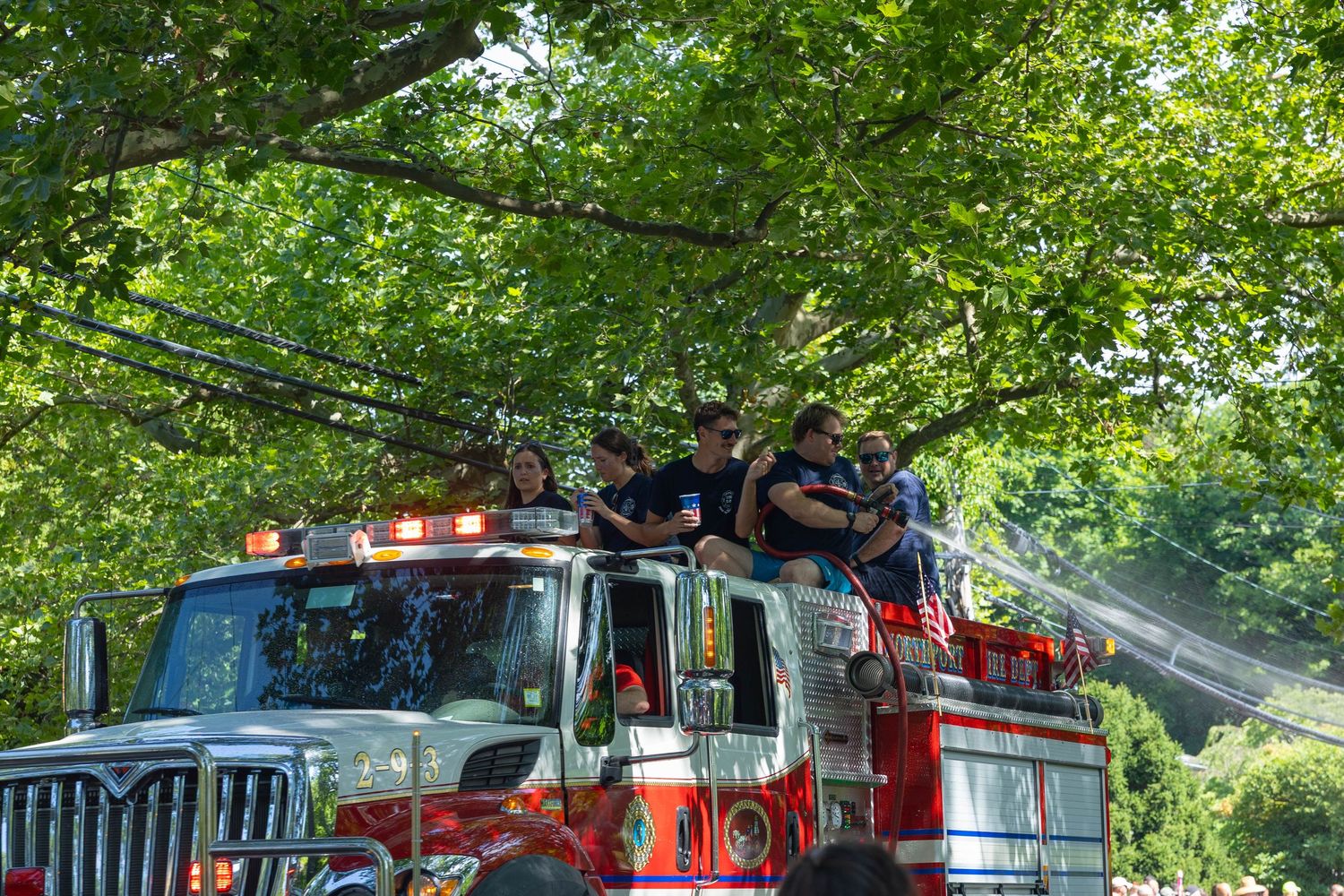Smiling firefighters ride on top of red International fire truck during a sunny community parade.