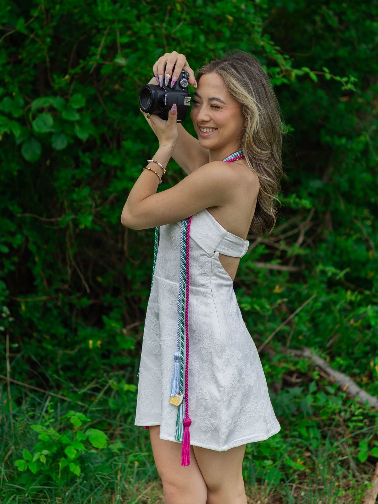 Smiling young woman in white dress holding a camera up to photograph outdoors in greenery.