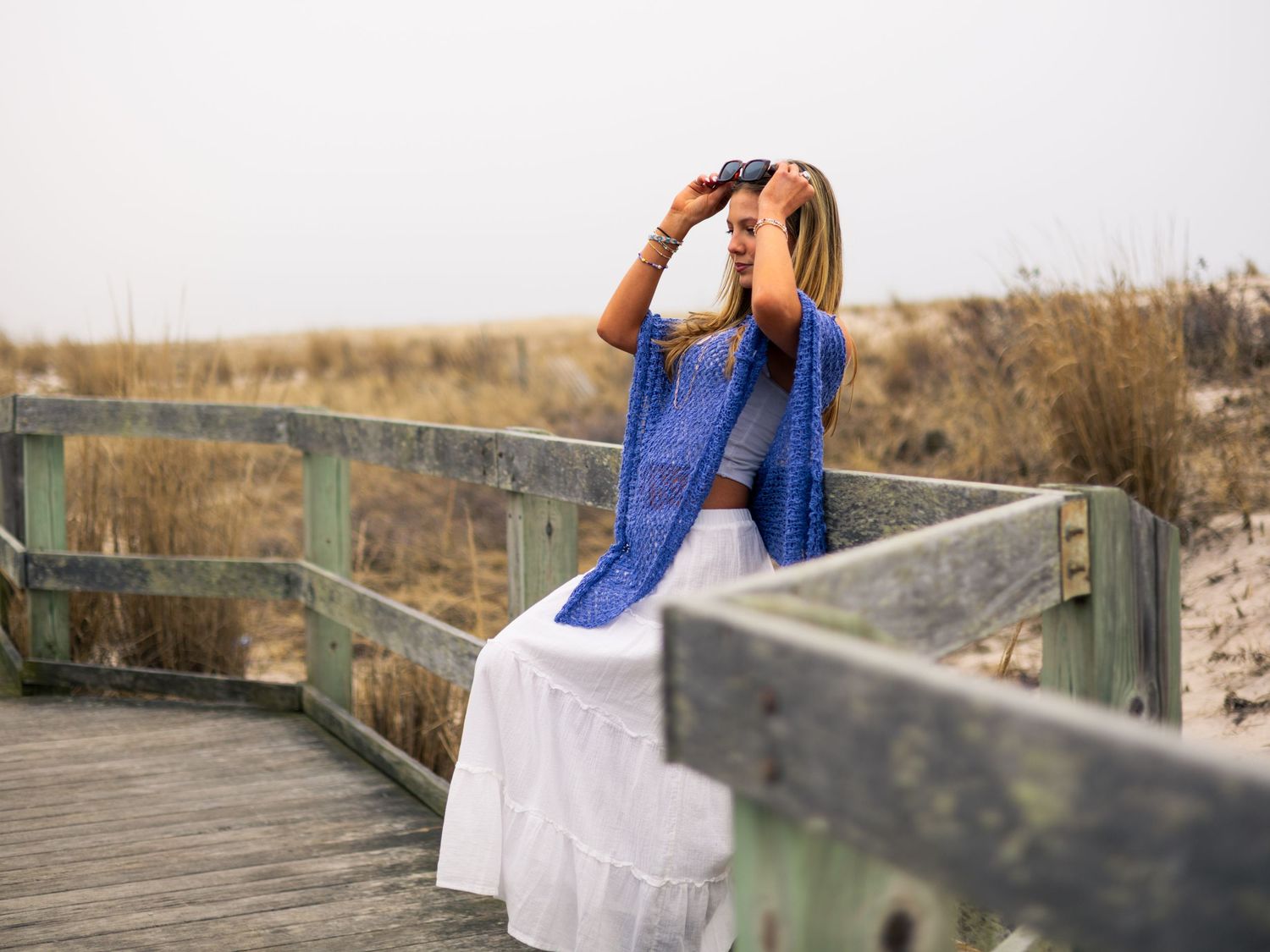 Woman in white skirt and blue scarf sitting on wooden boardwalk, looking through binoculars at a sandy beach.