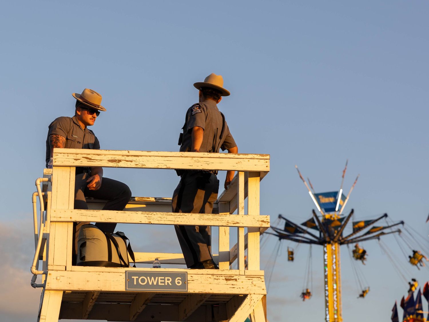 Two park rangers in campaign hats stand on Tower 6 platform at a fairground with amusement rides visible at dusk.