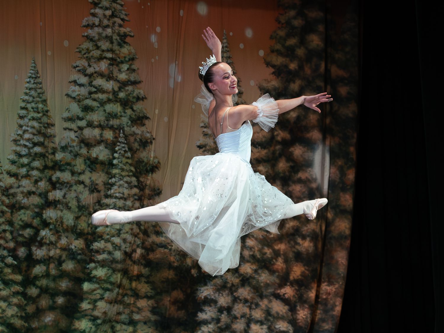Ballerina in white dress leaping gracefully on stage with evergreen tree backdrop during a Nutcracker ballet performance.