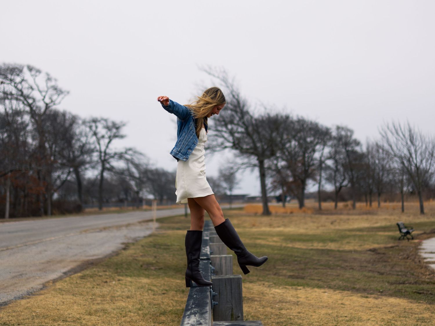 Young woman in a denim jacket, white skirt and black boots balances on a roadside guardrail beside a grassy field and bare trees.