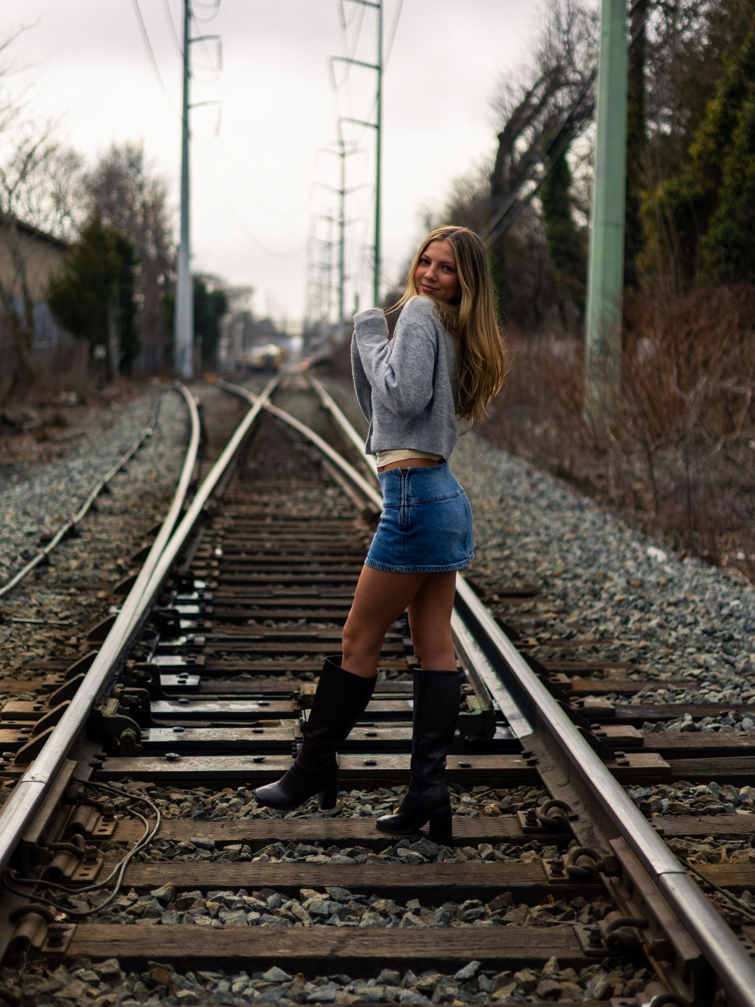 Young woman with long blonde hair poses on railroad tracks in a gray sweater, denim shorts and black knee-high boots on a cloudy day.