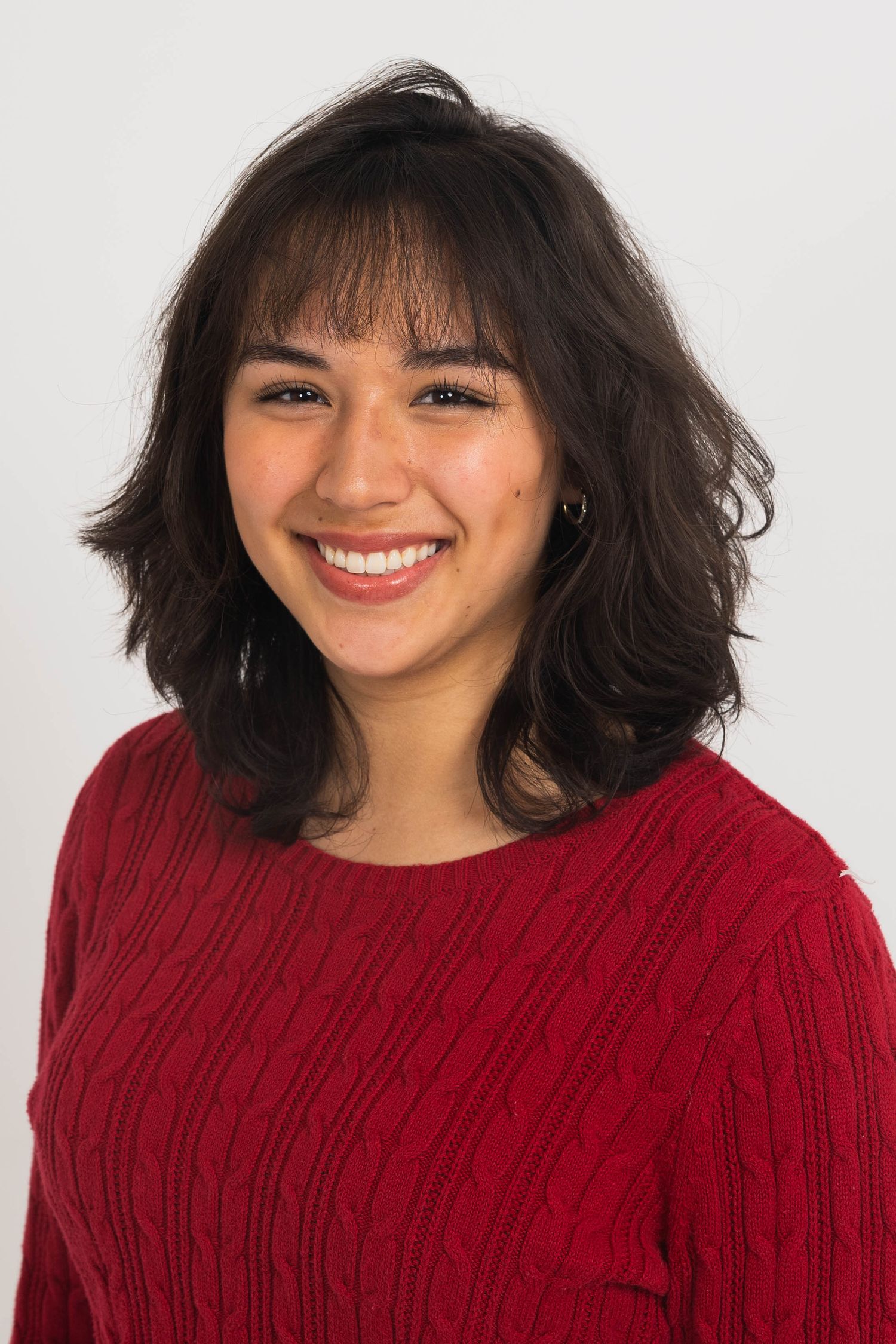 Smiling young woman with shoulder-length wavy dark hair and bangs wearing a red cable-knit sweater against a plain light background.