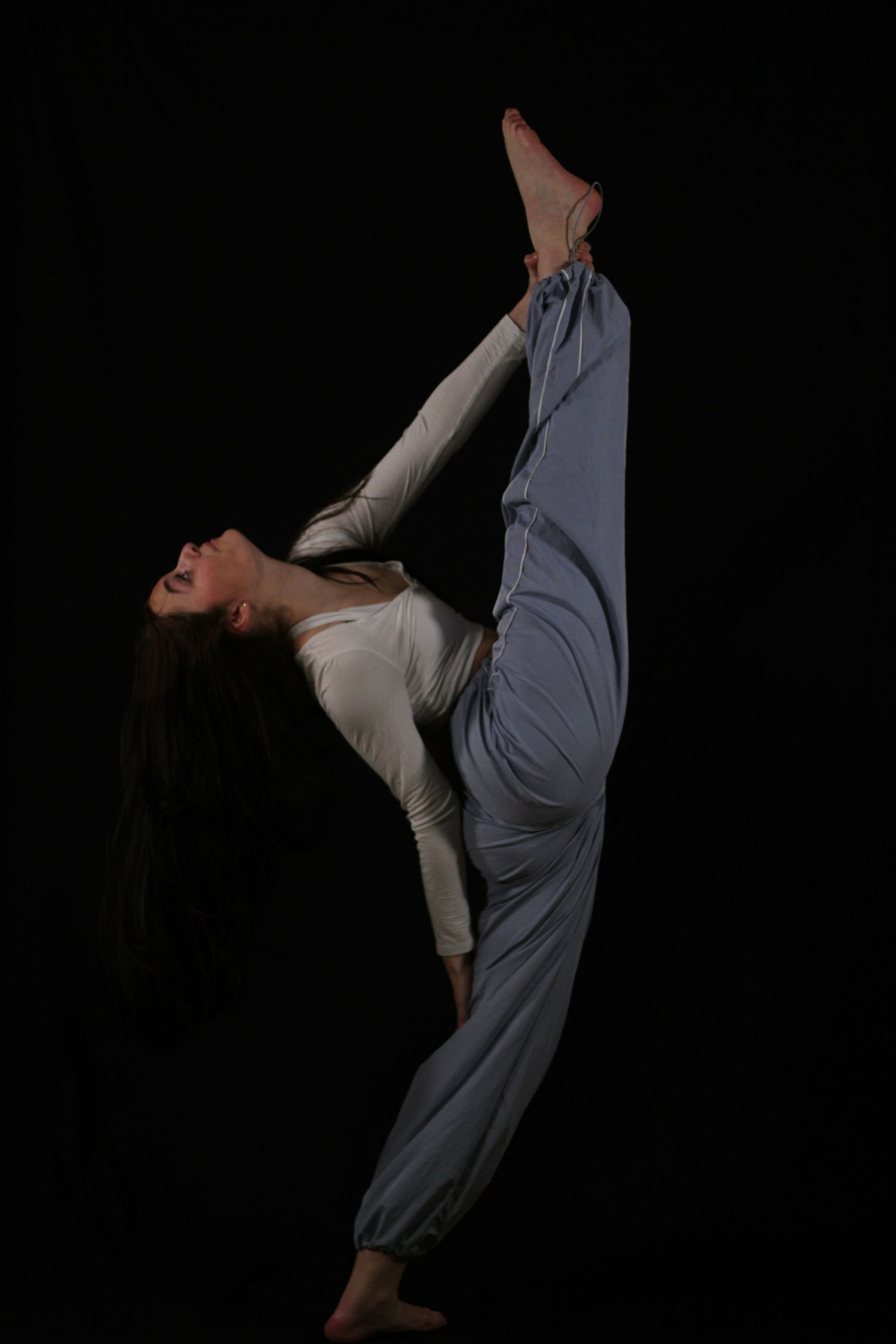 Dancer in a white top and gray harem pants performs a vertical standing split with long dark hair flowing against a black background.