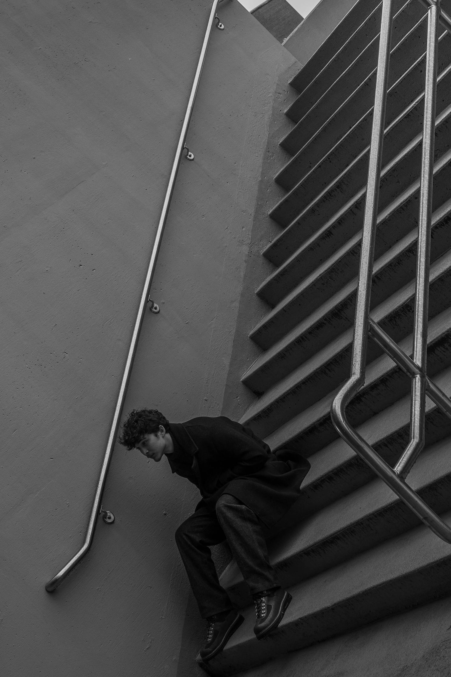 Black and white photo of a man in a dark coat and boots crouching on concrete stairs beside a metal handrail.