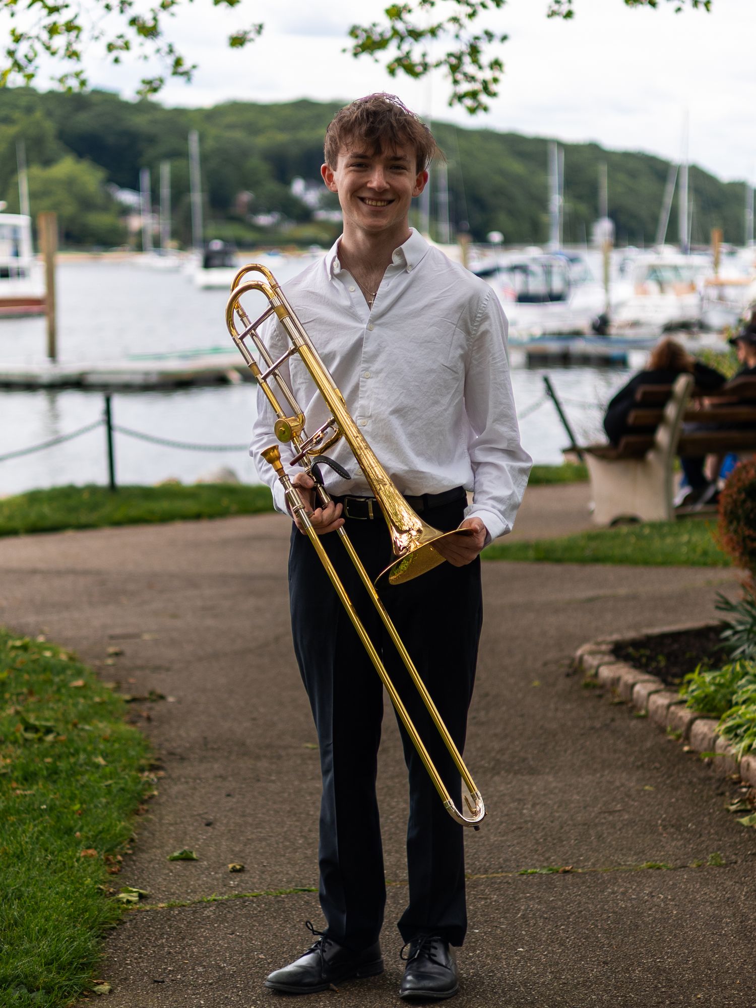 Young man smiling and holding a trombone outdoors near a marina on a sunny day.