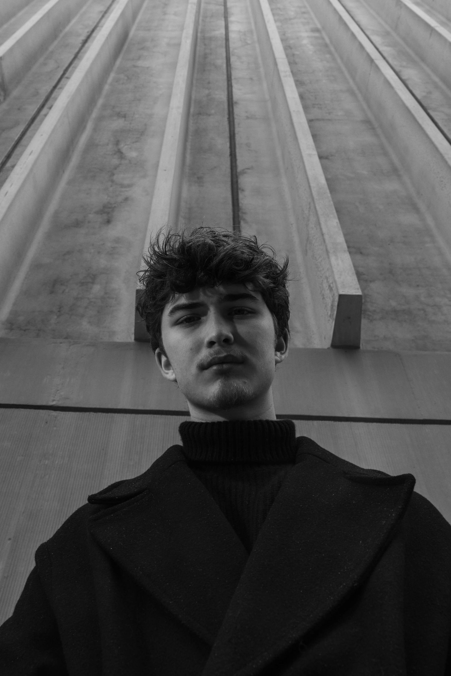 Black and white low-angle portrait of a young man with tousled hair in a dark wool coat and turtleneck against a textured concrete wall.