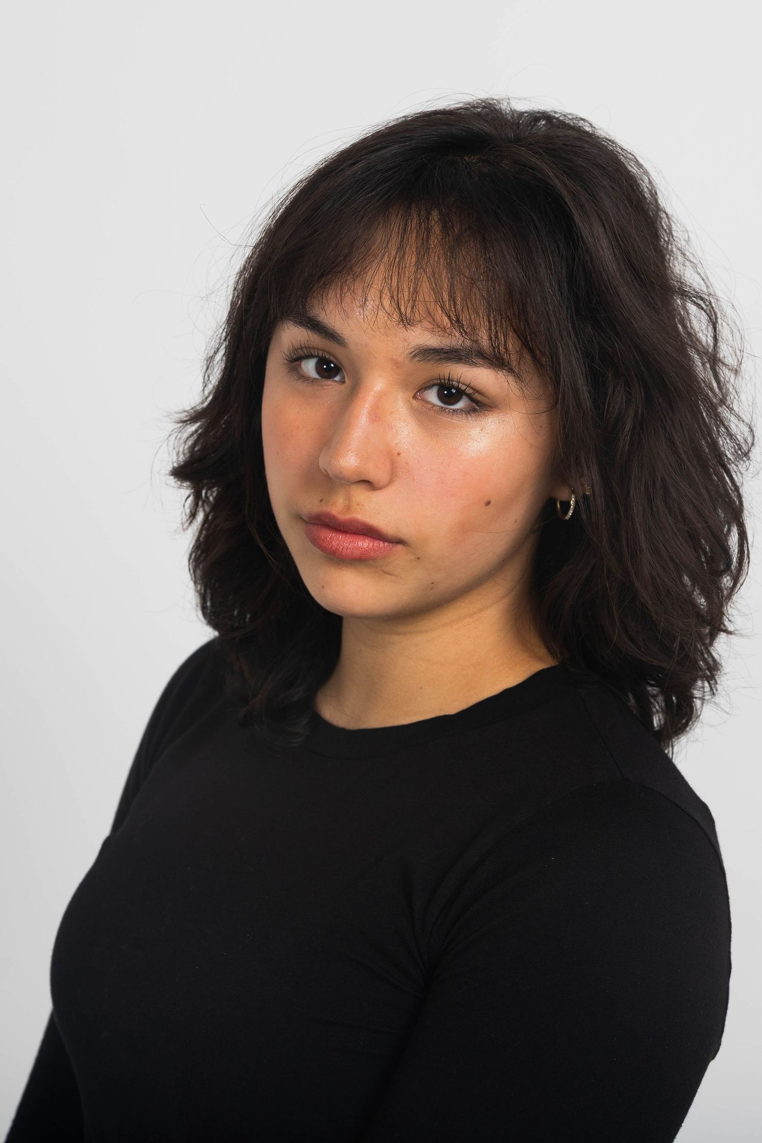 Young woman with dark wavy shoulder-length hair and bangs wearing a black long-sleeve top, posed against a plain white studio background.