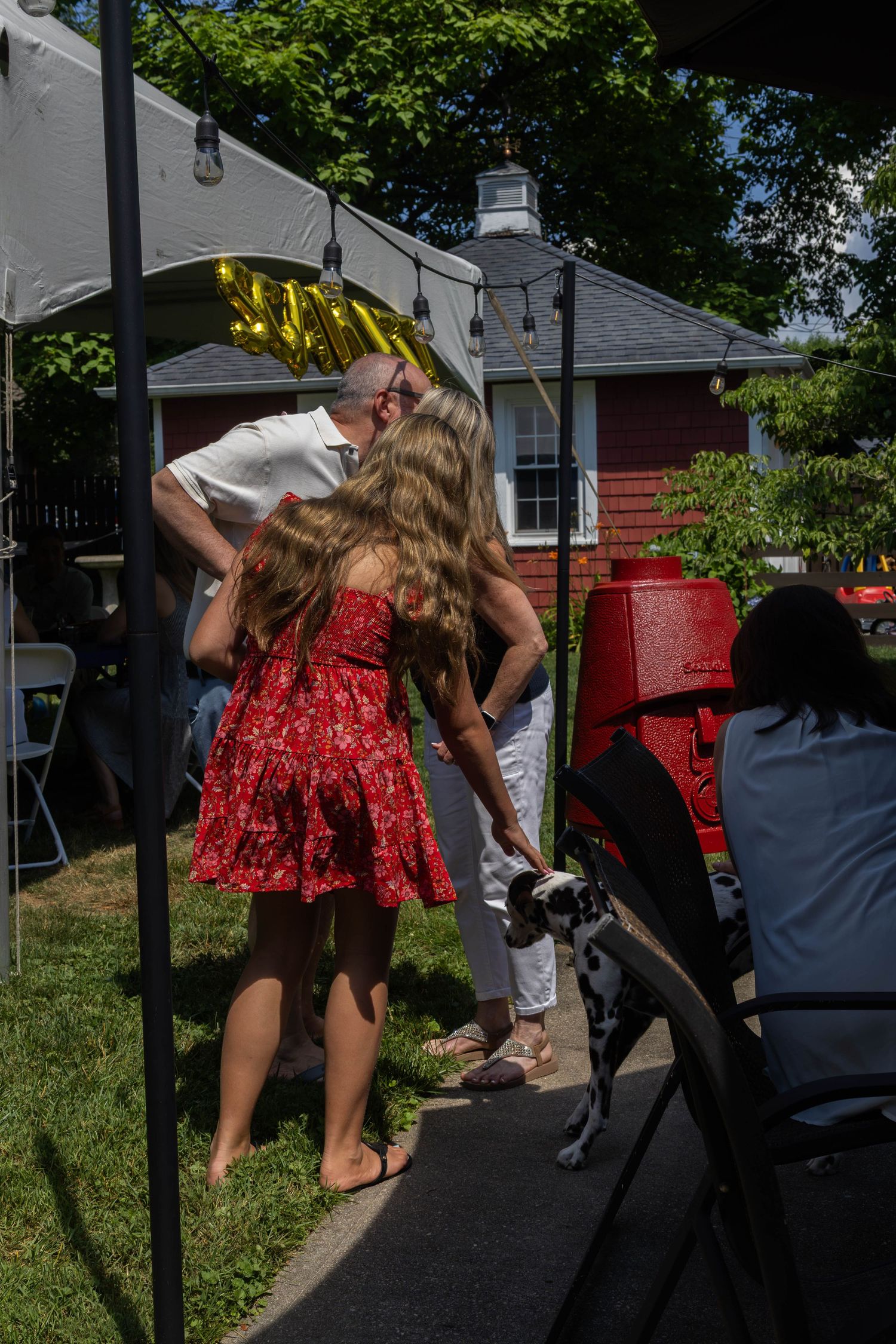 Young girl in red floral dress pets a dog at an outdoor backyard graduation celebration.