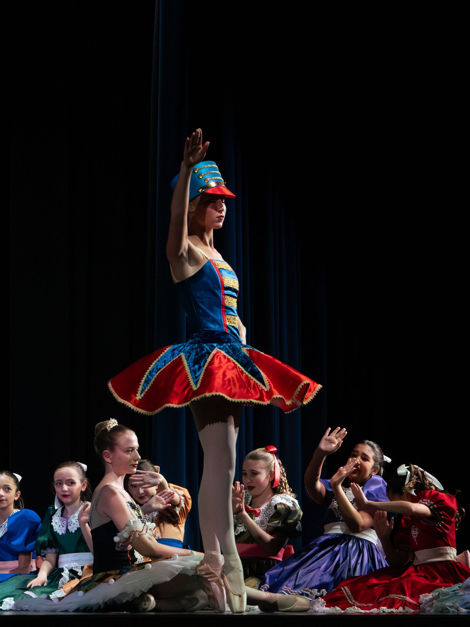 A ballerina in a blue and red tutu and hat performs on stage while young dancers sit behind her.