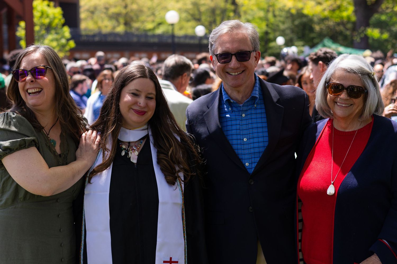 A graduate in cap and gown poses with three smiling family members outdoors at a sunny college commencement ceremony.