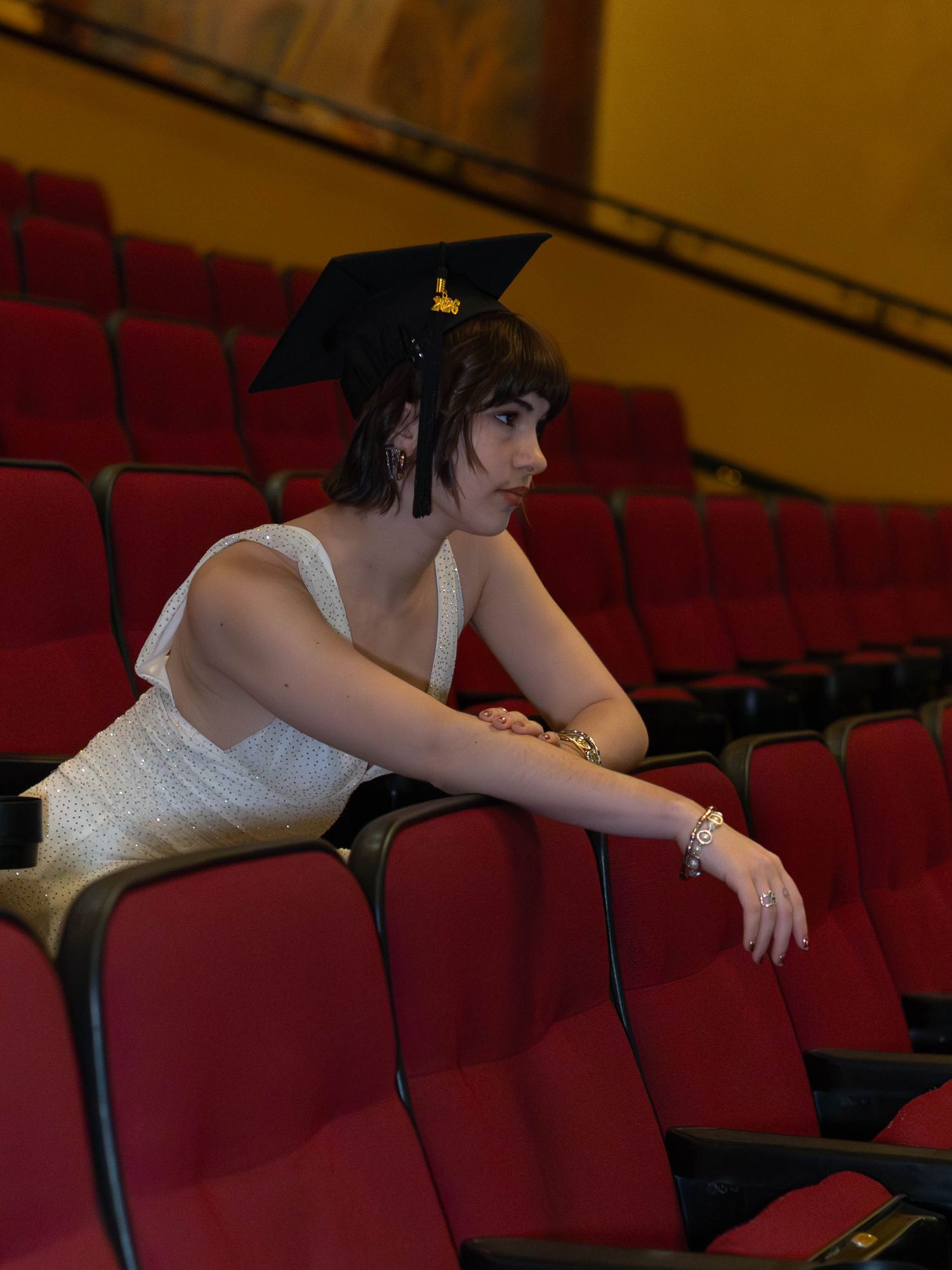 A graduate in cap and white dress sits among red theater seats, looking thoughtful.