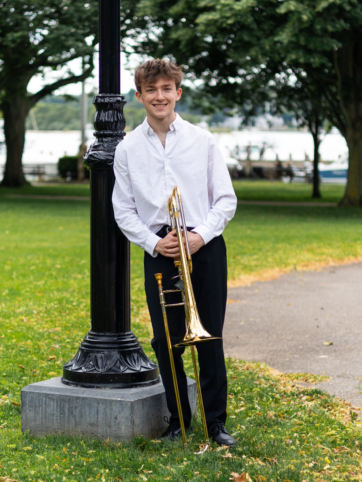 Young musician smiling outdoors, holding a trombone while leaning against a black lamppost in a green park.