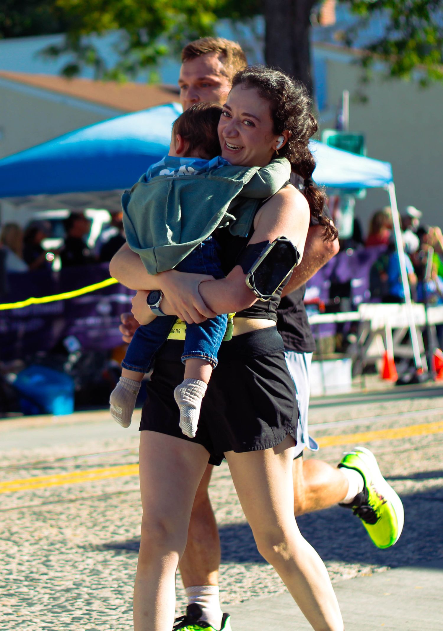 Smiling woman carrying a child while running in an outdoor race event on a sunny day.