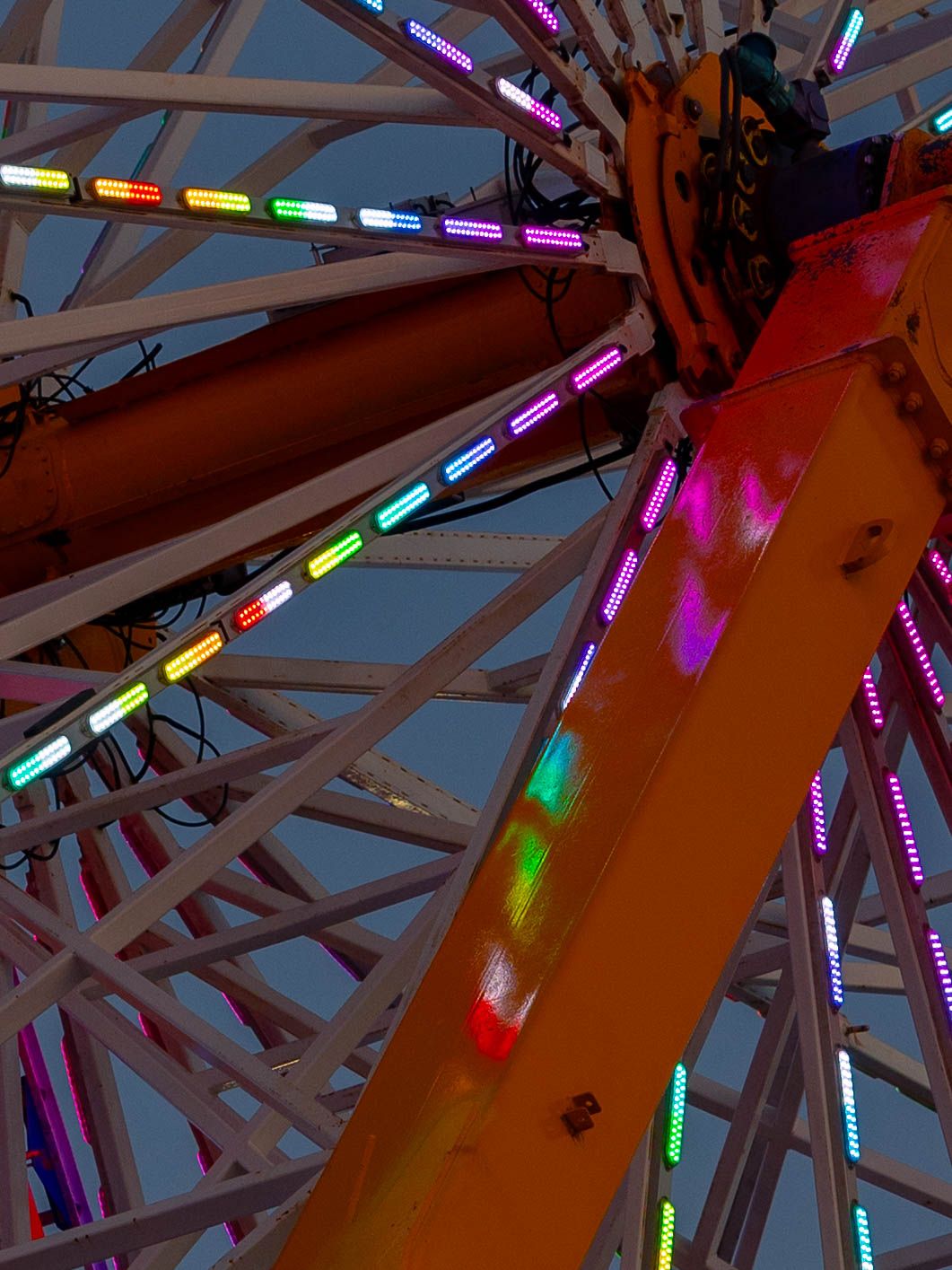 Colorful LED lights illuminate a Ferris wheel structure against a dusk sky at an amusement park or carnival.
