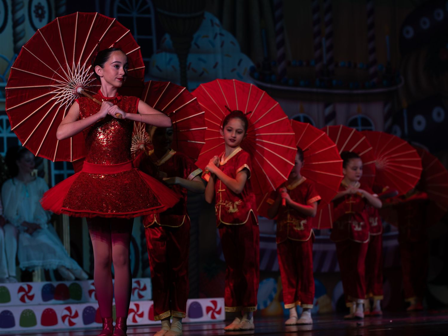 Young dancers in red costumes perform a Chinese dance with red parasols on a theatrical stage.