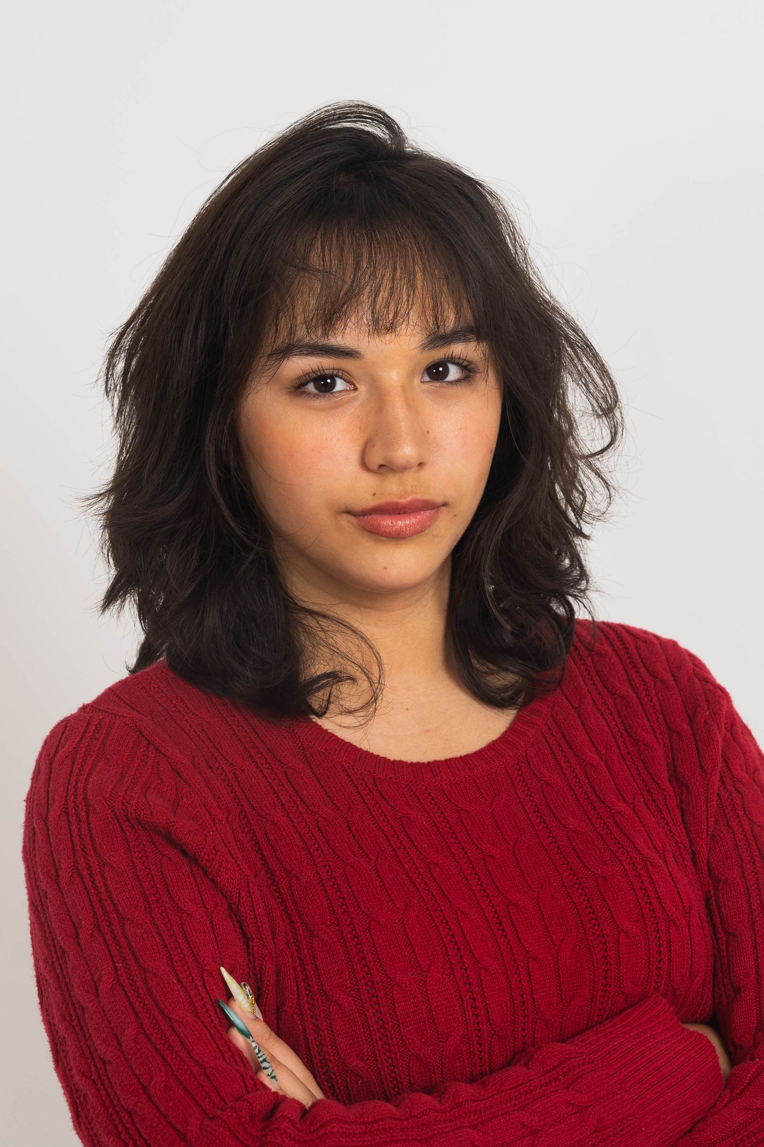 Young woman with wavy dark hair and bangs poses in a red cable-knit sweater with arms crossed against a plain light background.