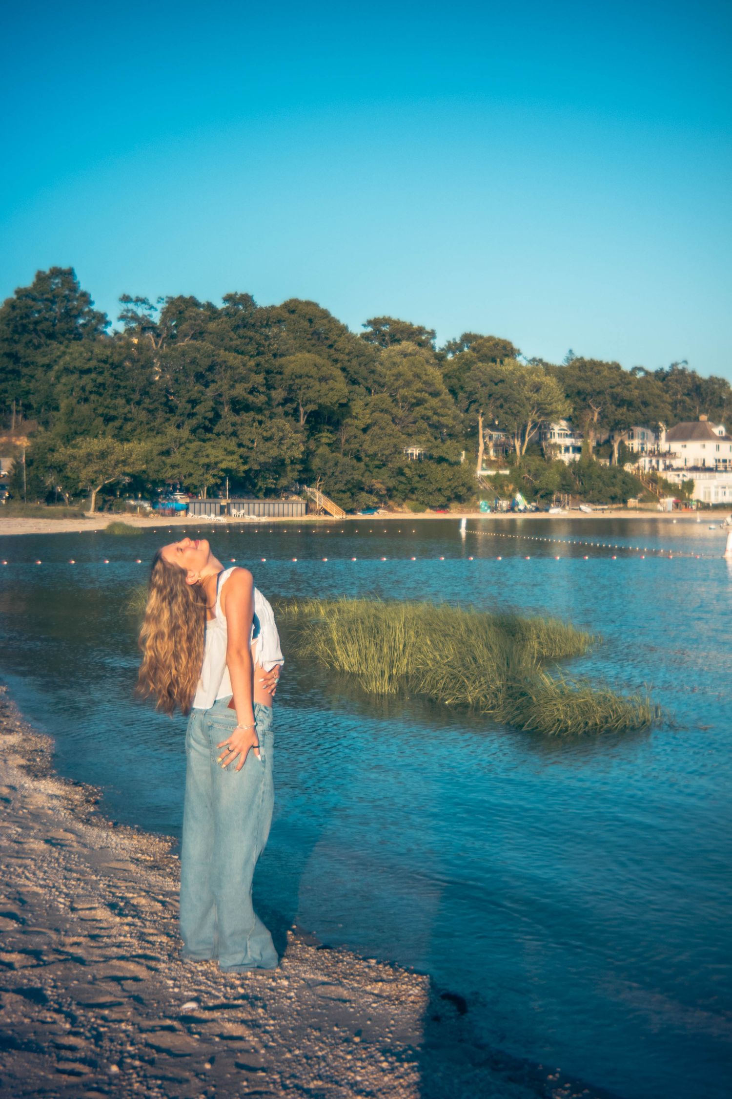 Woman with long hair tilts her head back while standing on a pebbly shore beside calm water, marsh grass, and tree-lined coastal homes.