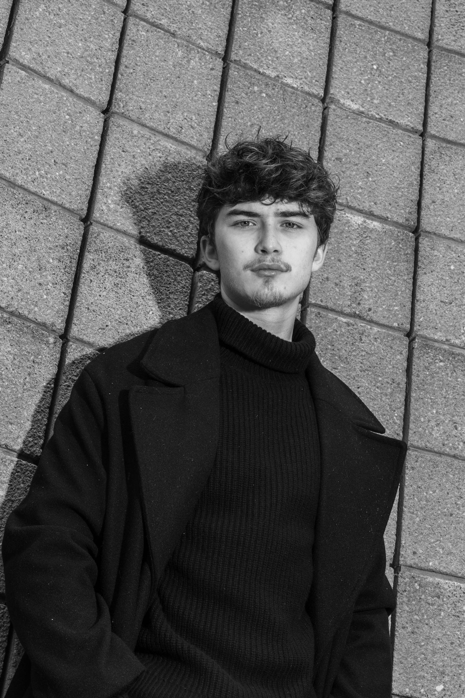 Black and white portrait of a young man with curly hair in a dark coat and turtleneck, leaning against a textured concrete block wall.