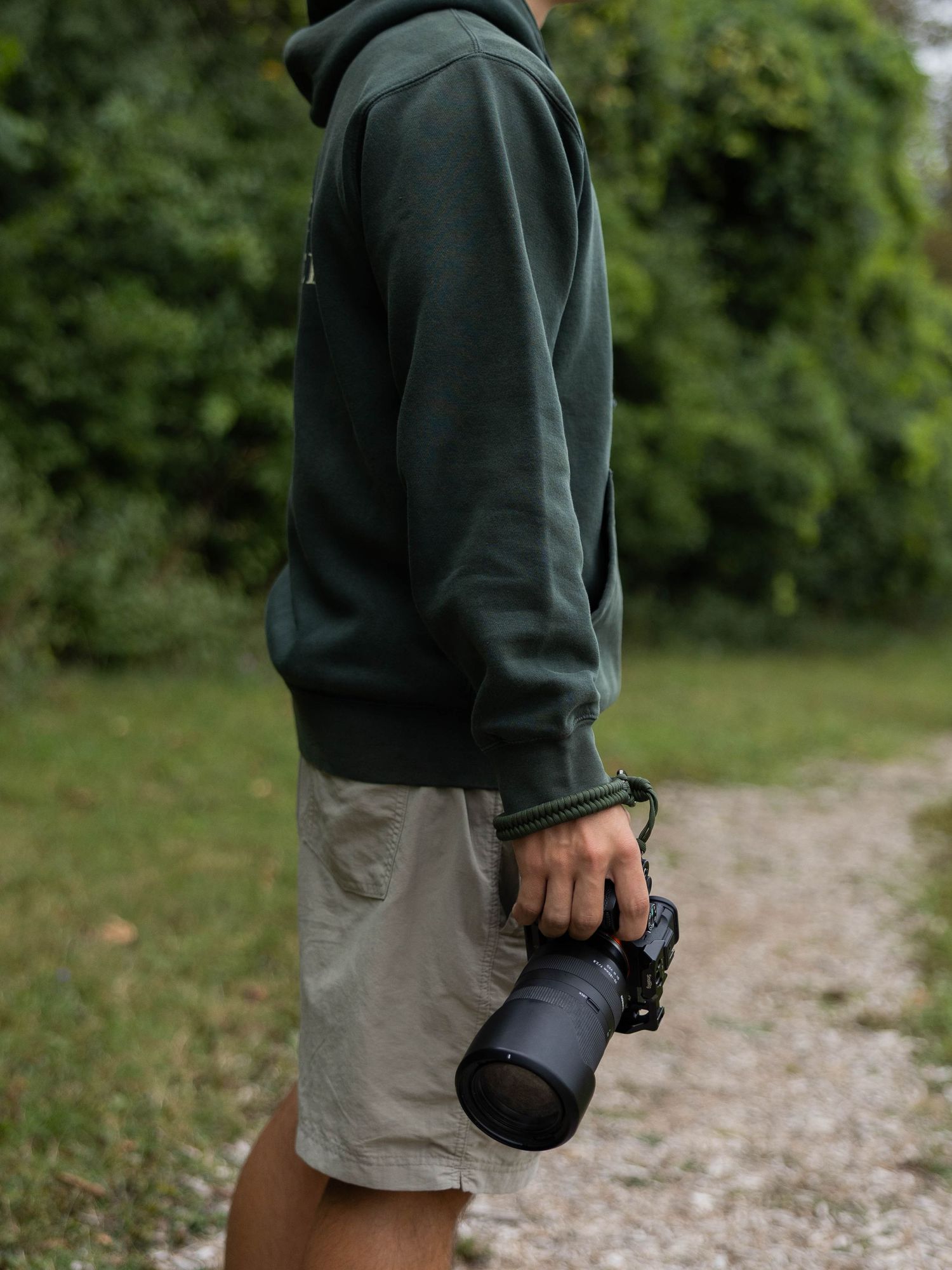 Person in green hoodie and khaki shorts holding a DSLR camera with telephoto lens outdoors.