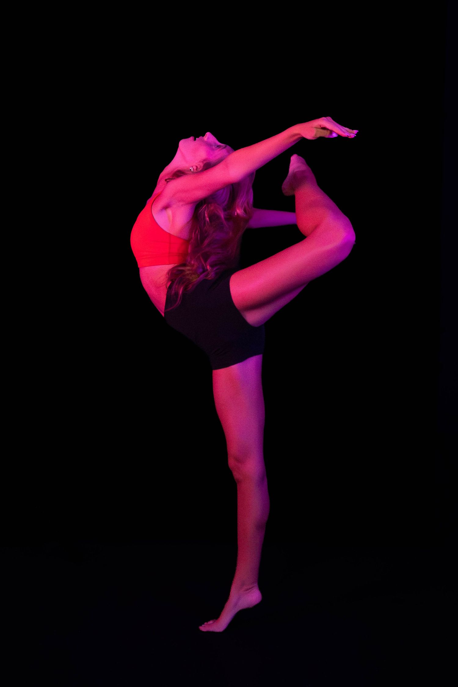 Dancer in a red top and black shorts performs a standing split with a deep backbend, lit in vivid pink and red against a black background.