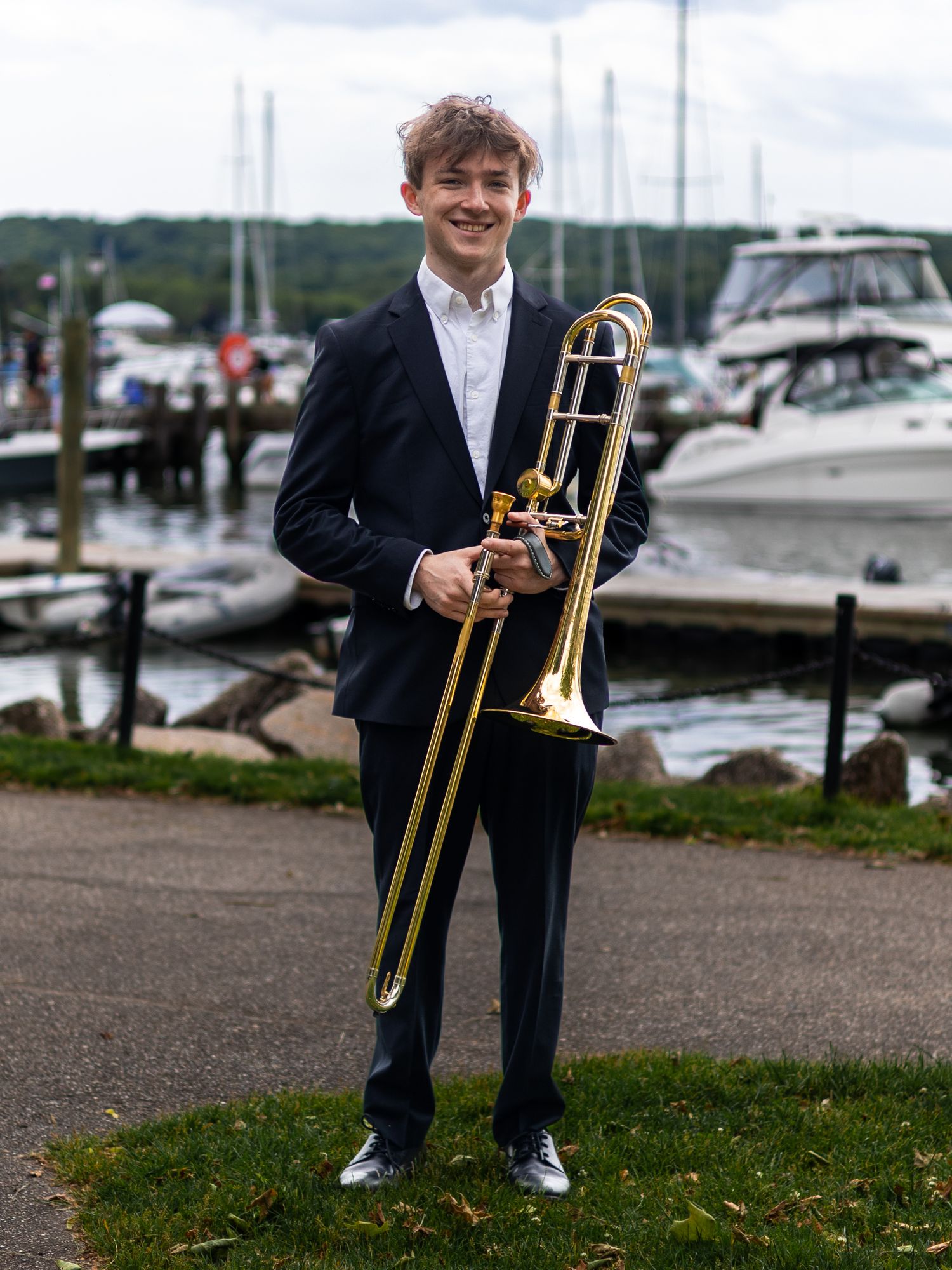 Smiling young man in navy suit holding trombone at marina with boats in background.