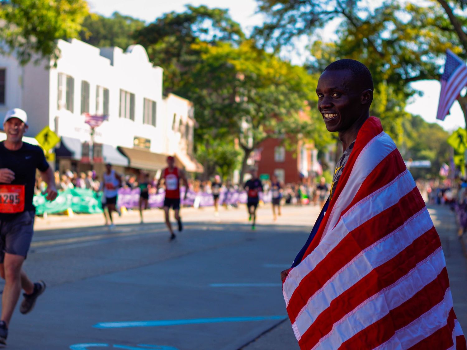 Cow Harbor Race winner wrapped in American flag smiles at camera during outdoor road race on sunny day.