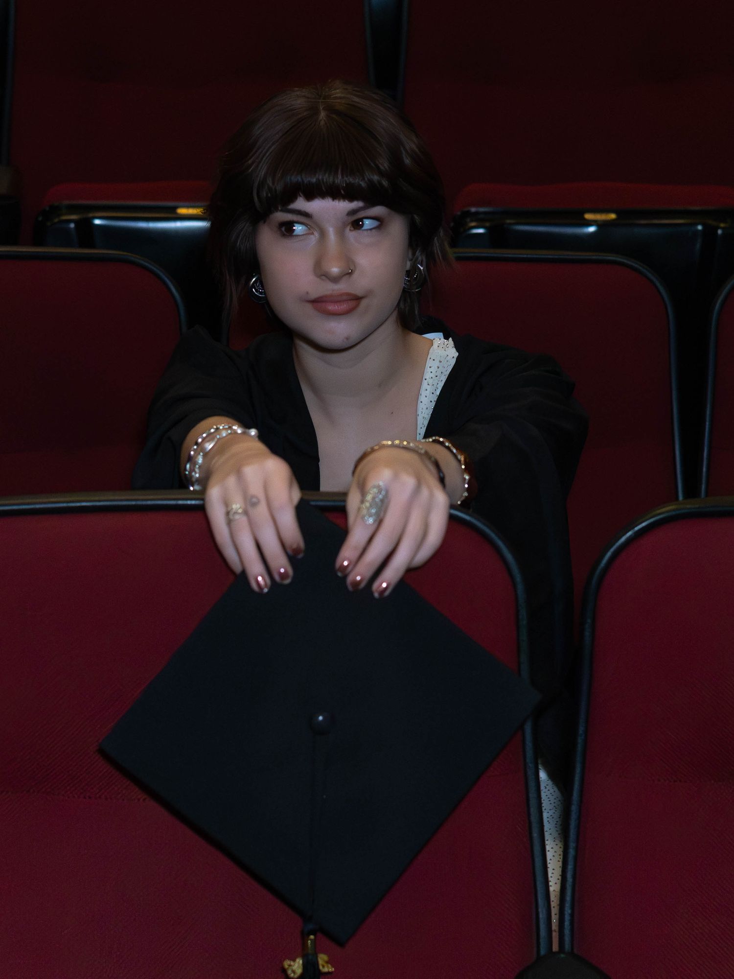 Young woman in black graduation gown holds mortarboard cap while seated in red auditorium chairs.