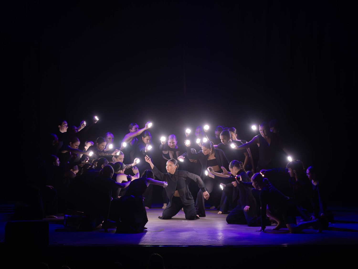 Performers in black costumes hold glowing lights on a dark stage during a dramatic group dance performance.