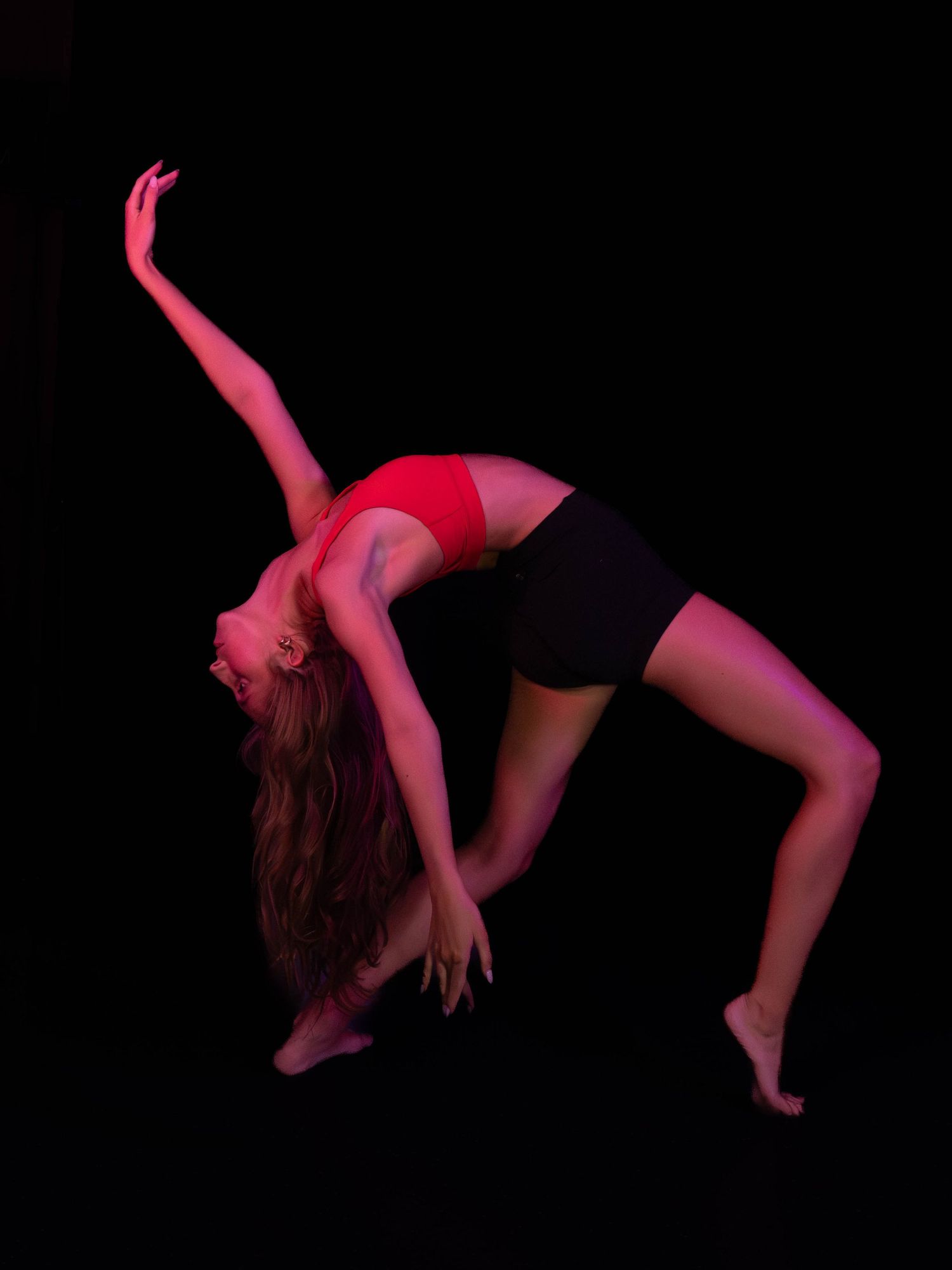 A dancer in a red top and black shorts arches backward in a dramatic pose, lit by moody red stage lighting against a black background.