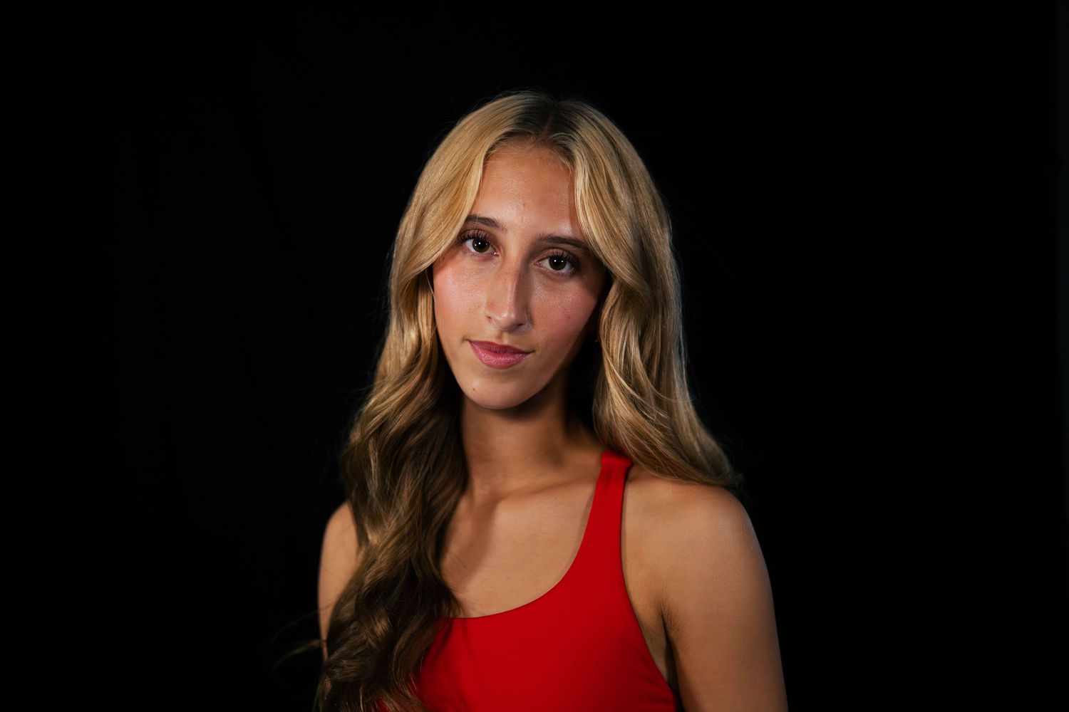 Young woman with long wavy blonde hair wearing a red sleeveless top poses against a solid black background.