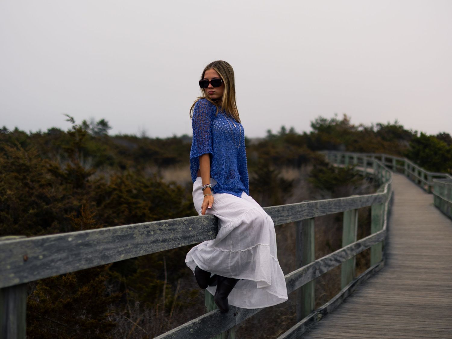 Woman in a blue knit top, white skirt and sunglasses sits on a wooden boardwalk railing surrounded by coastal shrubs at dusk.