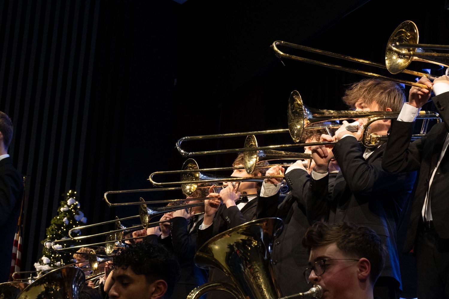 A row of trombone players perform together during a school band concert on a dark stage.