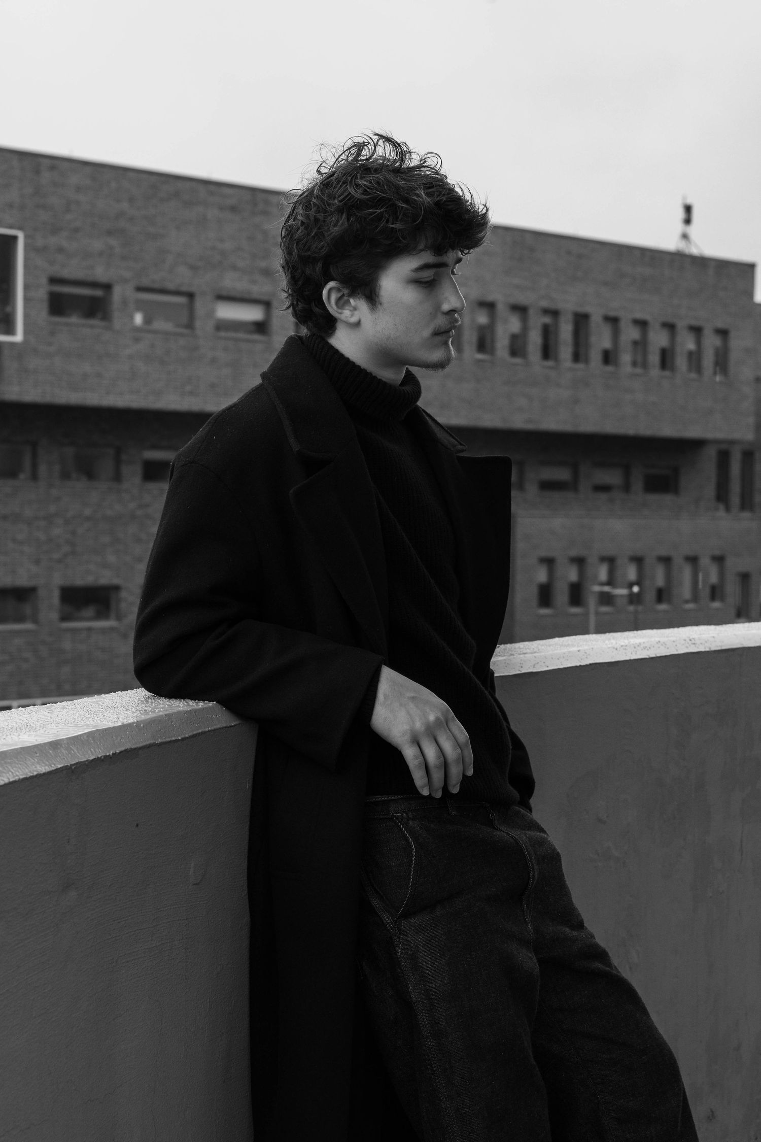 Black and white photo of a young man with curly hair in a long dark coat leaning on a rooftop ledge with a brick building behind.