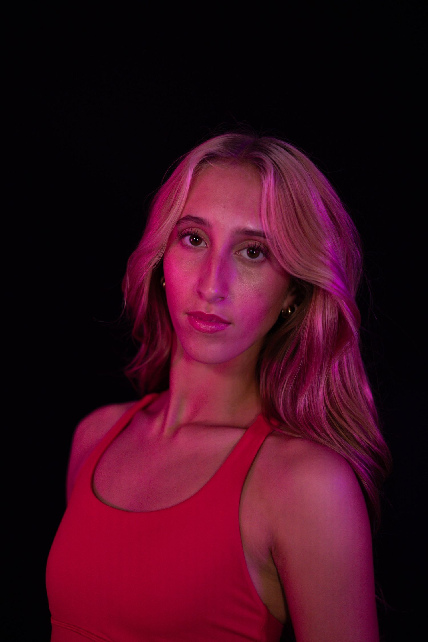 Portrait of a young woman with long wavy blonde hair in a red tank top, lit by vivid pink and magenta light against a dark background.