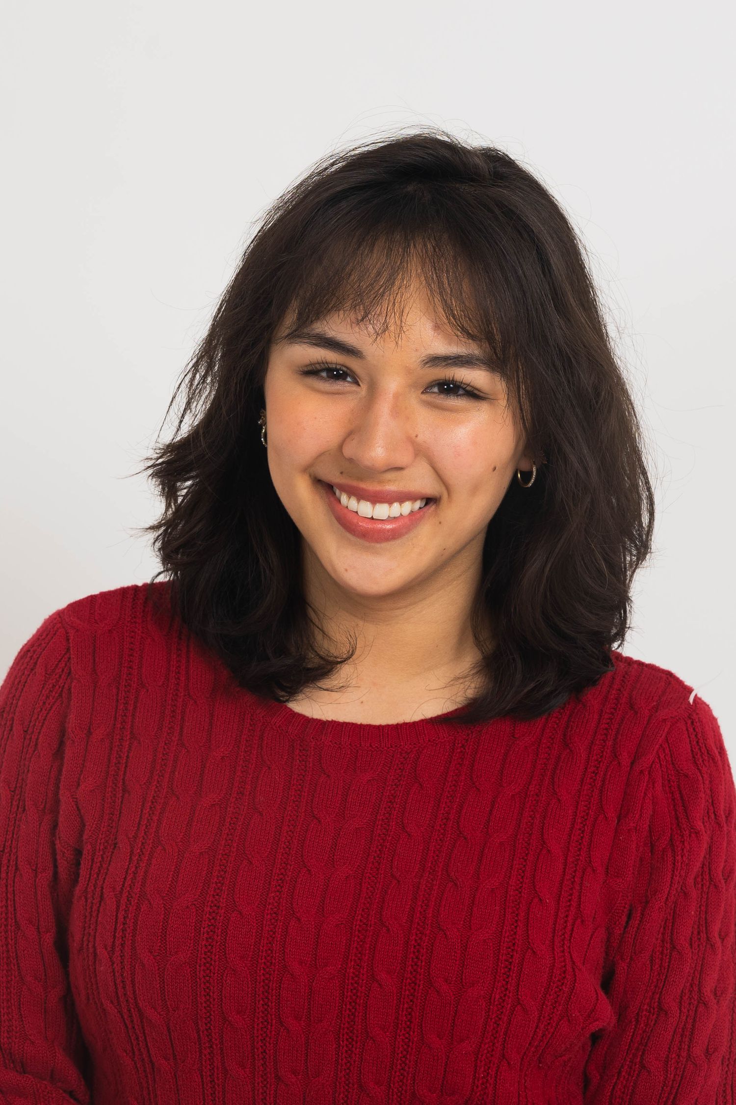 Smiling young woman with shoulder-length wavy dark hair and bangs wearing a red cable-knit sweater against a plain white background.