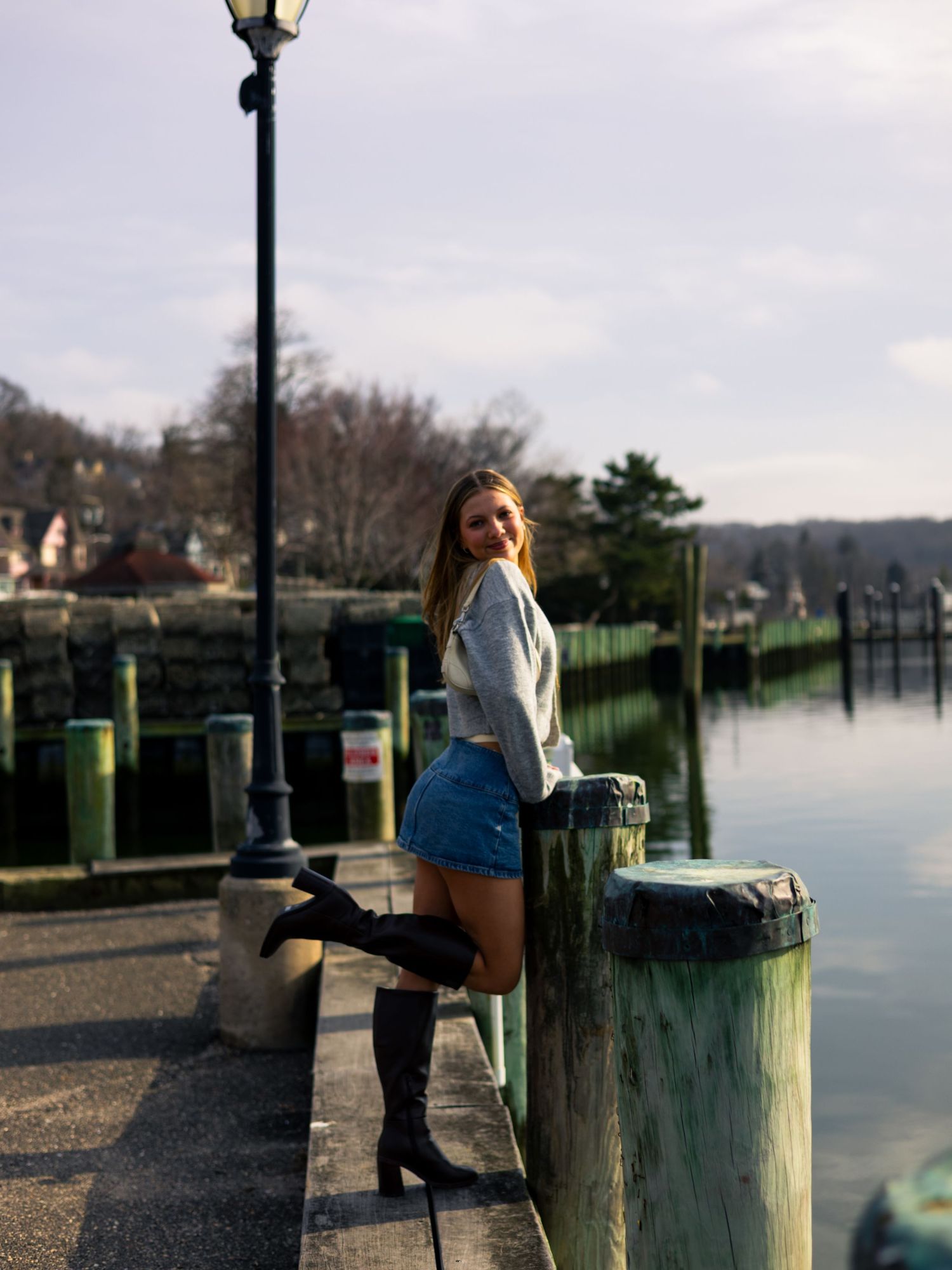 Young woman in denim skirt and black knee-high boots posing at a waterfront dock near a lamp post.
