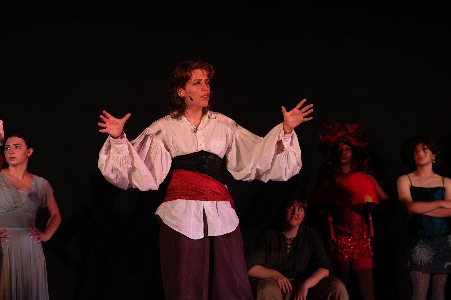A female performer in pirate costume gestures dramatically on stage with ensemble cast behind her.