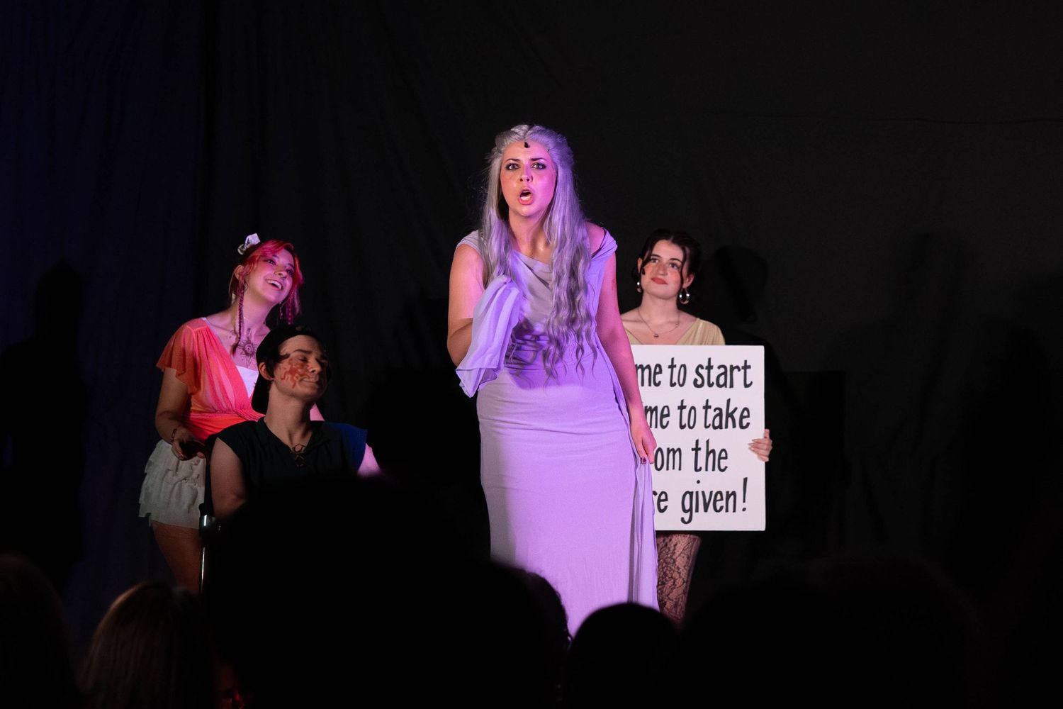 Performers on stage in costumes, one holding a sign, during a theatrical production under dramatic lighting.