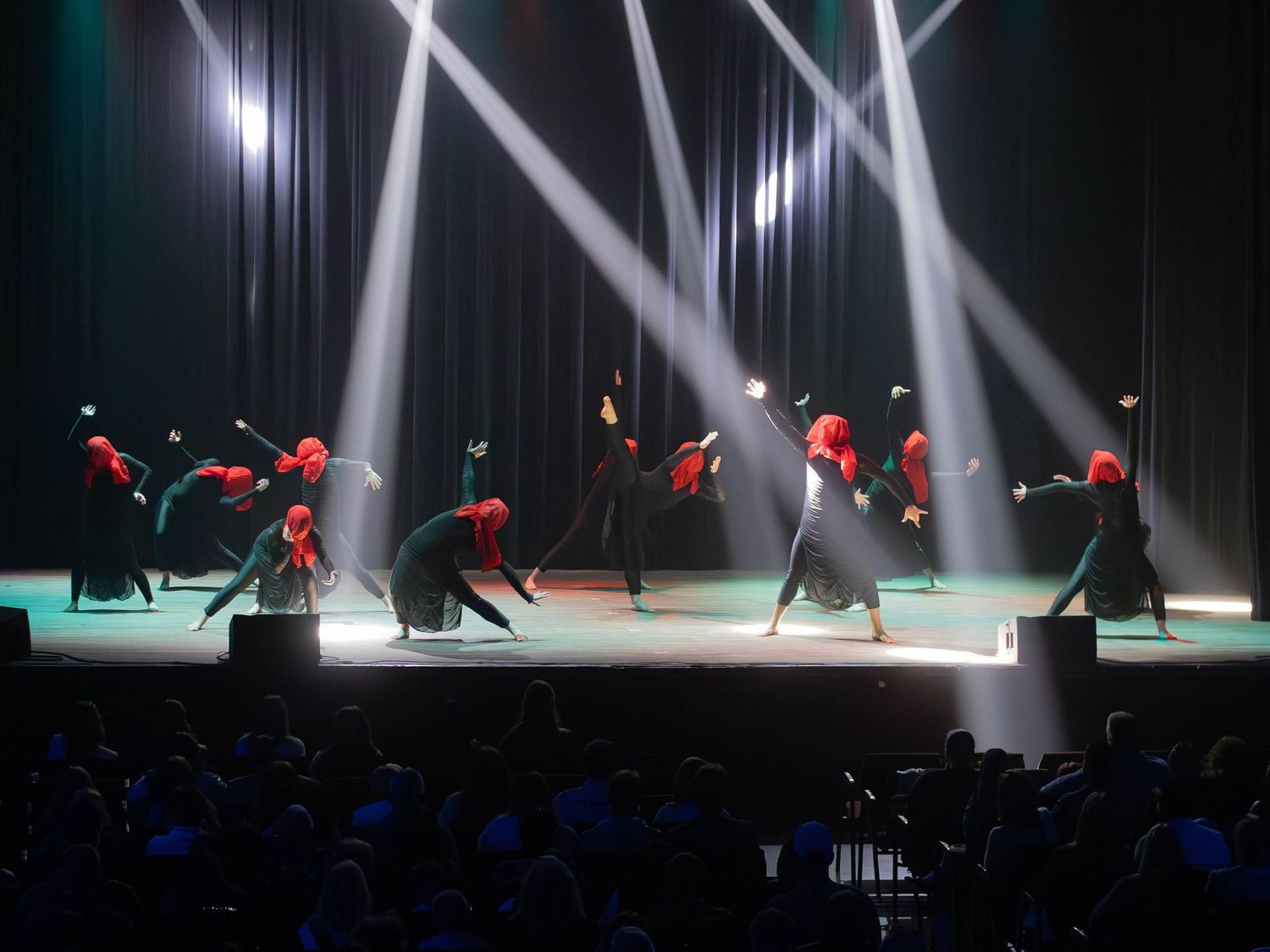 Dancers in red masks and black costumes perform on a dramatic stage lit by bright white spotlights.